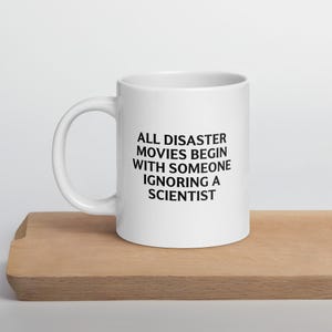 May include: White ceramic mug with the text "ALL DISASTER MOVIES BEGIN WITH SOMEONE IGNORING A SCIENTIST" in black. The mug is on a wooden surface.
