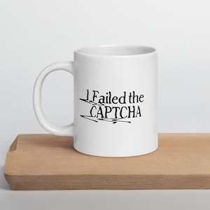 May include: White ceramic mug with the text "I Failed the CAPTCHA" in black, stylized font. The mug is cylindrical with a curved handle. The mug is sitting on a wooden surface.