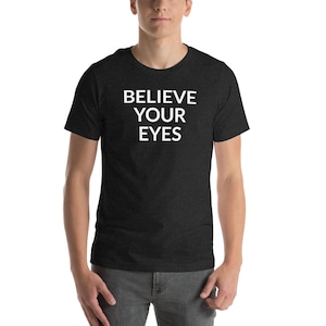 Puede incluir: Camiseta gris oscuro con el texto blanco "BELIEVE YOUR EYES". La camiseta es de manga corta y tiene cuello redondo. La persona lleva vaqueros grises.