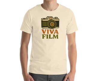 May include: Cream-colored t-shirt featuring a vintage camera design with a sunflower lens. The words "VIVA FILM" are printed below the camera in brown and green. The shirt is short-sleeved and has a crew neck.