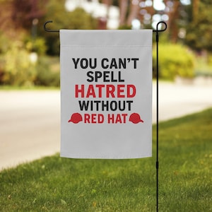 May include: A white garden flag with the text "YOU CAN'T SPELL HATRED WITHOUT RED HAT" in black and red. The flag includes two red baseball cap graphics. The flag is hanging on a black metal pole in a grassy yard.