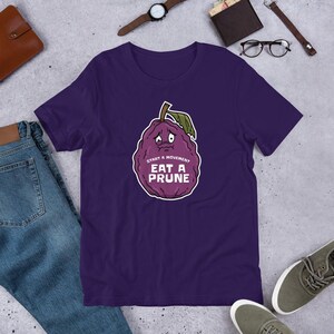 May include: Purple t-shirt with a cartoon prune with a sad face and the text "Start a movement Eat a Prune".
