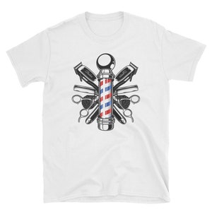 Puede incluir: Una camiseta blanca con una ilustraci&oacute;n vintage de un poste de barbero rodeado de herramientas de barbero, incluyendo tijeras, navajas y cepillos. El poste de barbero es rojo, blanco y azul con una base negra.