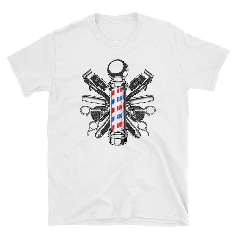Puede incluir: Una camiseta blanca con una ilustraci&oacute;n vintage de un poste de barbero rodeado de herramientas de barbero, incluyendo tijeras, navajas y cepillos. El poste de barbero es rojo, blanco y azul con una base negra.