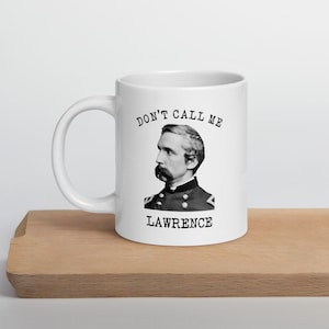 May include: White ceramic mug featuring a black and white portrait of a man with a mustache and the text "DON'T CALL ME LAWRENCE". The mug has a standard handle and is likely used for hot beverages.