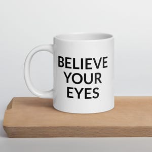 Puede incluir: Taza de cerámica blanca con la frase "BELIEVE YOUR EYES" impresa en letras negras en negrita. La taza tiene un asa curva y está sobre una superficie de madera. Una taza sencilla para café o té.