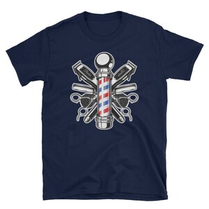 Puede incluir: Camiseta azul marino con una ilustraci&oacute;n vintage de un poste de barbero rodeado de herramientas de barbero cruzadas. El poste de barbero es rojo, blanco y azul con una base plateada. Las herramientas incluyen cortadoras, navajas y un peine.