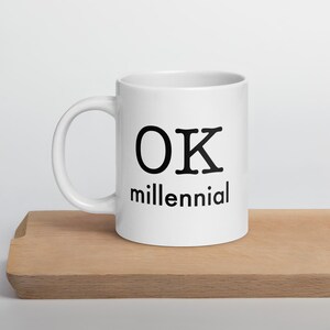 May include: White ceramic mug with the text "OK millennial" in black. The mug has a curved handle and sits on a wooden surface. The mug is a standard size for coffee or tea.
