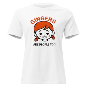 May include: White t-shirt with a cartoon illustration of a girl with red hair and the text "GINGERS ARE PEOPLE TOO" in orange. The graphic includes heart-shaped details on the face. The shirt has short sleeves and a crew neck.