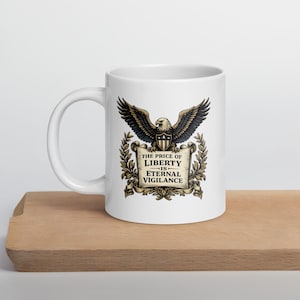 May include: White ceramic mug featuring a detailed illustration of an eagle with outstretched wings, a shield, and a banner that reads "THE PRICE OF LIBERTY IS ETERNAL VIGILANCE." The design is in shades of brown, gold, and black.