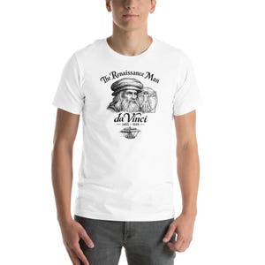 Puede incluir: Camiseta blanca con una ilustración en blanco y negro de Leonardo da Vinci. El texto dice "The Renaissance Man" y "da Vinci 1452-1519". El diseño incluye un retrato, un dibujo anatómico y un barco.