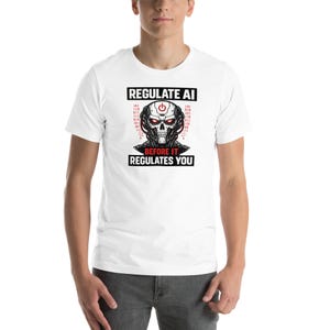 May include: White t-shirt with a graphic of a robotic skull in red and black, with binary code elements. The text reads "REGULATE AI" above the skull and "BEFORE IT REGULATES YOU" below. The shirt is short-sleeved.