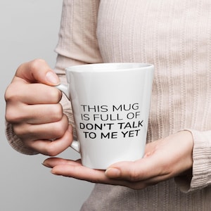 May include: A white ceramic mug with a tapered shape and a handle on the left side. The mug features black printed text that reads: THIS MUG IS FULL OF DON'T TALK TO ME YET. The design is centered on the front of the smooth, glossy white surface.