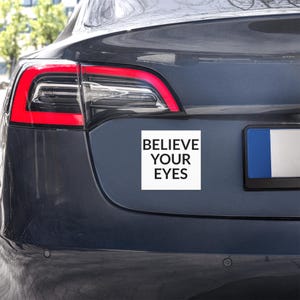 Puede incluir: Un imán de coche cuadrado blanco con el texto "BELIEVE YOUR EYES" en negro, fijado en la parte trasera de un coche gris oscuro. El piloto trasero del coche es visible, con un contorno rojo.