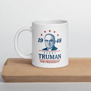 May include: White ceramic mug featuring a portrait of Harry Truman, the text "HARRY TRUMAN FOR PRESIDENT," and the numbers "1948." The design includes red stars and blue text. The mug is on a wooden surface.