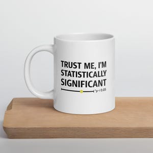 Può includere: Tazza in ceramica bianca con la scritta nera "TRUST ME, I'M STATISTICALLY SIGNIFICANT". Sotto il testo c'è una linea nera con un punto giallo e "*p<0.05". La tazza è su una superficie di legno.