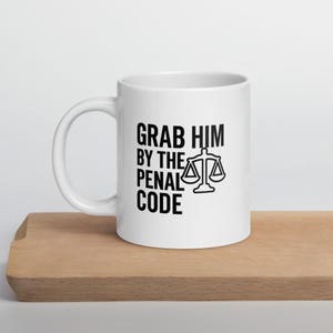 May include: White ceramic mug with black text that reads "Grab Him By The Penal Code" and a scale of justice graphic.