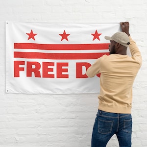 May include: White flag with three red stars and horizontal red stripes, with the words "FREE D." in large red letters. The flag is being held up against a white brick wall.