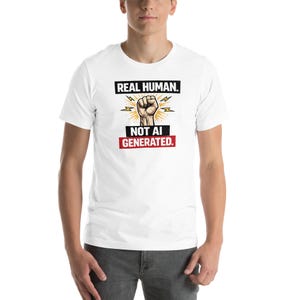 May include: White t-shirt with the text "REAL HUMAN." above a graphic of a clenched fist with lightning bolts, and the text "NOT AI GENERATED." in a red box. The shirt is short-sleeved.
