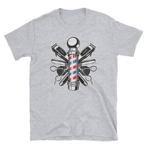 Puede incluir: Una camiseta gris con una ilustraci&oacute;n vintage de un poste de barbero rodeado de herramientas de barbero, incluyendo tijeras, navajas y peines. El poste de barbero es rojo, blanco y azul.