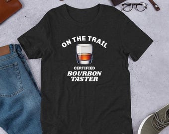 May include: A dark heather grey t-shirt with a white graphic design. The graphic features a glass of bourbon and the text "On the Trail Certified Bourbon Taster".
