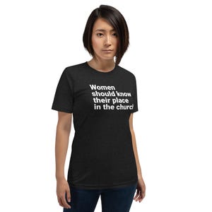 May include: A dark heather gray short-sleeve t-shirt featuring white text printed on the chest that reads: Women should know their place in the church. The garment has a crew neckline and a standard fit, constructed from a soft, heathered fabric.