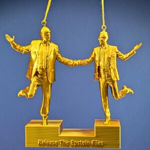 Puede incluir: Adorno dorado que representa dos figuras con trajes, tomadas de la mano, con los brazos extendidos. Las figuras están sobre plataformas separadas, con el texto "Release The Epstein Files" en la base. El adorno cuelga de un hilo dorado sobre un fondo azul.