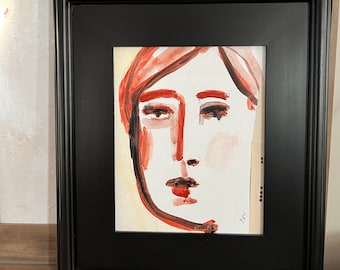 May include: Abstract face art in a black frame. The artwork features a minimalist style with bold red and black brushstrokes defining the facial features against a white background. The framed art is displayed on a wooden surface.