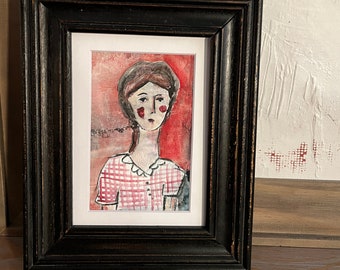 Op de afbeelding: Een ingelijste aquarel van een vrouw met een rood-wit geruit shirt en een rode achtergrond. De schildering zit in een zwarte, verweerde houten lijst met een witte passe-partout.