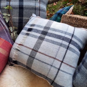 May include: Several square pillows with a plaid pattern in shades of gray, navy, and burgundy. The pillows have a fringed edge and are arranged outdoors. The pillows are made of a soft, woven fabric.