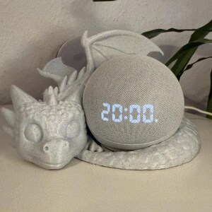 May include: A grey smart speaker displaying 20:00, set within a dragon-shaped holder. The holder is intricately designed with wings and a detailed dragon head, providing a decorative and fantasy-inspired setting.
