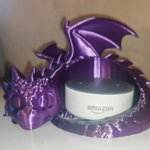 May include: A purple dragon-shaped smart speaker holder. The dragon has a detailed head, wings, and tail, cradling a white Amazon Echo Dot. The Amazon logo is visible on the speaker. The holder is made of a 3D-printed material.