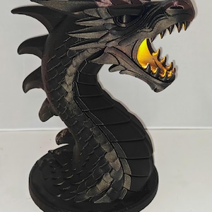 May include: A black dragon sculpture with an open mouth revealing a yellow glow. The dragon has detailed scales and a curved neck, resting on a round base. The sculpture's head is angled upwards, showcasing its sharp teeth and textured features.