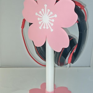 May include: A pink cherry blossom-shaped headphone stand with a white stem and a flower-shaped base. Black and red headphones are hanging on the stand. The stand is designed to hold headphones and keep them organised.