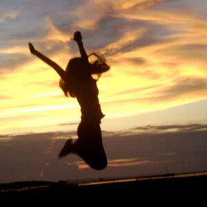 May include: Silhouette of a person jumping in the air with their arms raised against a bright orange and yellow sunset sky.