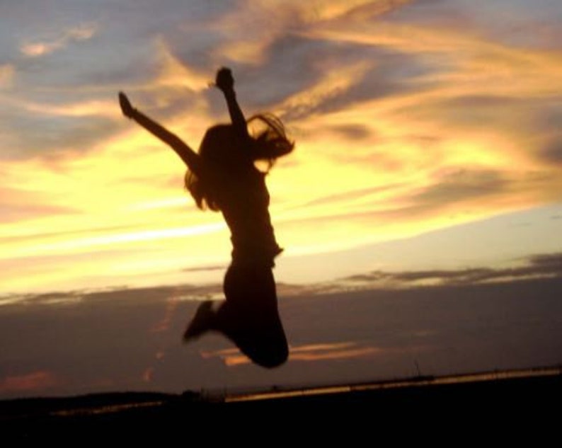 May include: Silhouette of a person jumping in the air with their arms raised against a bright orange and yellow sunset sky.