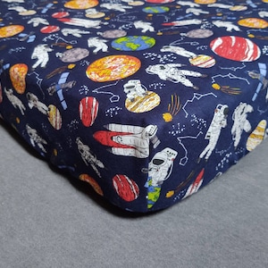Può includere: Un lenzuolo con angoli elasticizzato blu scuro con una stampa a tema spaziale che presenta astronauti, pianeti e stelle. La stampa è in tonalità di bianco, rosso, arancione e giallo.