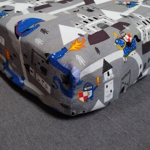 May include: A grey and white cot sheet with a repeating pattern of blue dragons, knights, and castles. The sheet features a whimsical design with a medieval theme.