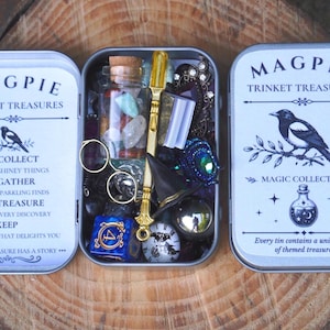 May include: A silver metal tin labelled Magpie Trinket Treasures Magic Collection containing a glass bottle with colourful stones, a gold-toned wand, a miniature black hat, a blue die with the number four, and various small decorative items.
