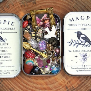 May include: A rectangular metal tin container with a hinged lid, featuring the text Magpie Trinket Treasures and a magpie illustration. The open centre compartment is filled with assorted small items including a miniature gold frame, a tiny spoon, beads, and a small glass bottle with a cork stopper.