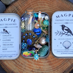 May include: A rectangular metal tin labelled Magpie Trinket Treasures Ocean Collection, filled with a variety of small items including a corked glass bottle, blue beads, a miniature bird, seashells, a turtle charm, and a vintage-style brooch.