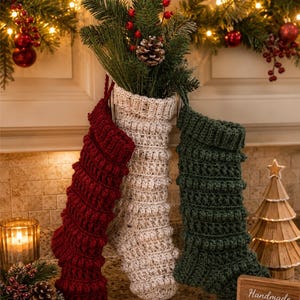 Puede incluir: Tres calcetines navideños de ganchillo en rojo oscuro, crema y verde oscuro, cada uno con un patrón texturizado de puntos de burbuja. Los calcetines se muestran con ramas de pino y piñas en su interior, creando un adorno festivo.
