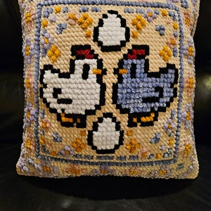Peut inclure: Un coussin beige avec un motif brodé coloré représentant deux poulets et des œufs. Les poulets sont blancs et bleus avec des crêtes rouges. Les œufs sont blancs.