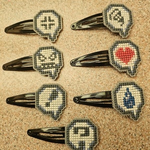 May include: Six black hair clips with different colored pixelated designs. The designs include a cross, a skull, a heart, a lightning bolt, a question mark, and a ghost.