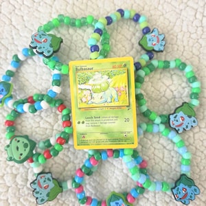 May include: A collection of colorful beaded bracelets featuring Bulbasaur charms and a Bulbasaur Pokémon card. The bracelets are made with green, blue, red, and white beads. The card is yellow and green.