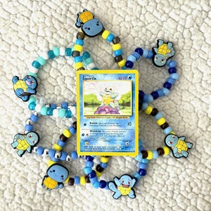 May include: A Squirtle Pokémon card is surrounded by beaded bracelets with Squirtle charms. The bracelets feature blue, yellow, and white beads. The card has a blue border and features a Squirtle illustration.