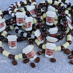 Puede incluir: Primer plano de varios brazaletes hechos con cuentas marrones, blancas y negras. Cada brazalete tiene un dije de una taza de café blanca con la palabra "Coffee" impresa en ella. Dispersos alrededor de los brazaletes hay granos de café marrones.