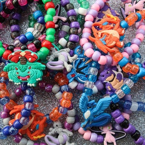 May include: A collection of colorful beaded bracelets with various Pokemon character charms. The bracelets are made with small, round beads in shades of blue, green, purple, pink, orange, and red. The charms include a green Venusaur, a blue Blastoise, a red Charizard, a purple Gengar, and a pink Jigglypuff.
