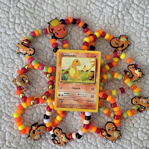 May include: A Charmander Pokémon trading card is surrounded by a beaded bracelet with Charmander charms. The bracelet features orange, yellow, black, and red beads. The card shows a yellow Charmander with the text "Charmander" and "50 HP".