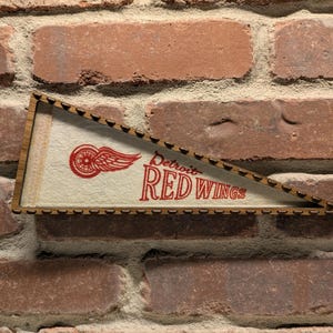May include: A wooden pennant with a white background and red lettering. The pennant reads "Detroit Red Wings" and features a red wing with a black wheel in the center.